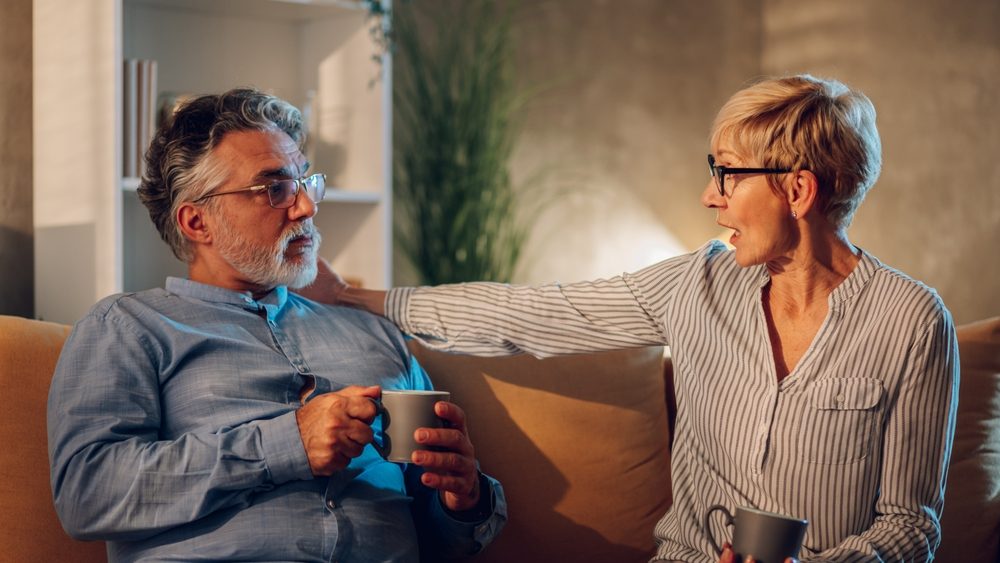 Portrait of a senior couple drinking coffee while sitting on a sofa at home and looking at each other while having a serious conversation. Modern interior. Copy space.