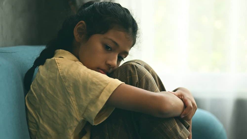 Unhappy girl portrait, Sad child sitting on sofa at home, Upset girl hugging knees alone, Concept of lonely girl and kid with trouble and violence