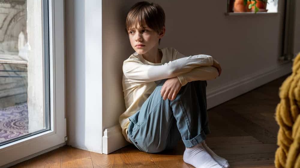 Kid psychological trauma. Lonely child boy sadly look to window hug knees sits on floor at home alone. Upset offended child thinking about family problems, bad relationship, failures with school peers