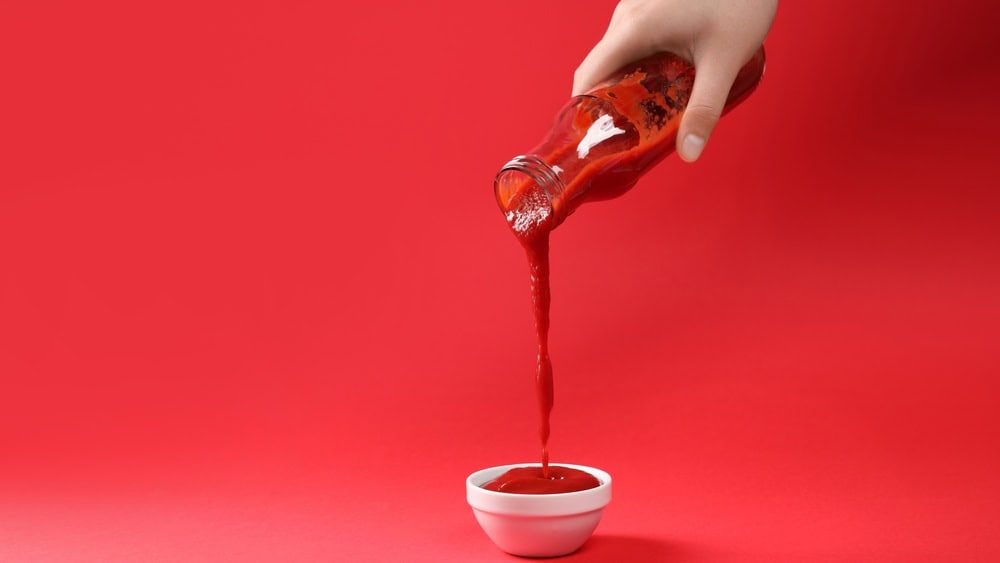 Woman pouring tasty ketchup from bottle into bowl on red background, closeup. Space for text