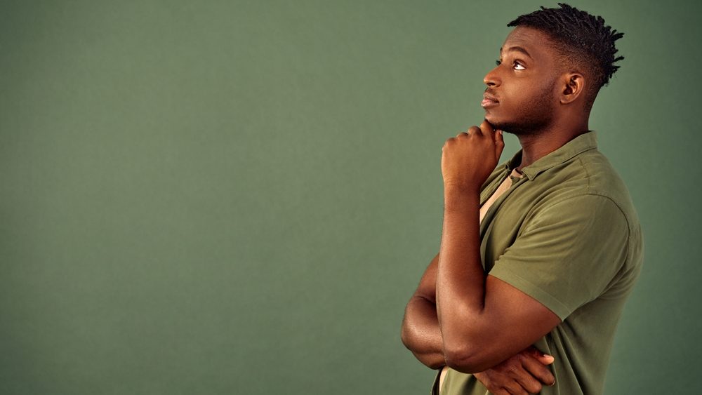 Lost in thoughts. Side view of handsome african american young man with pensive facial expression holding hand near chin while standing in studio. Isolated over green background with copy space.