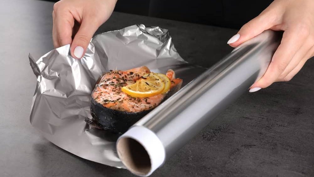 Woman wrapping tasty salmon with lemon and spices in aluminum foil at grey textured table, closeup