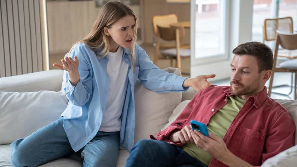 Indignant, offended, hysterical woman screams at husband sitting on sofa with cellphone. Couple family quarrel conflict misunderstanding discord. Indifferent man ignores dissatisfied wife's questions.