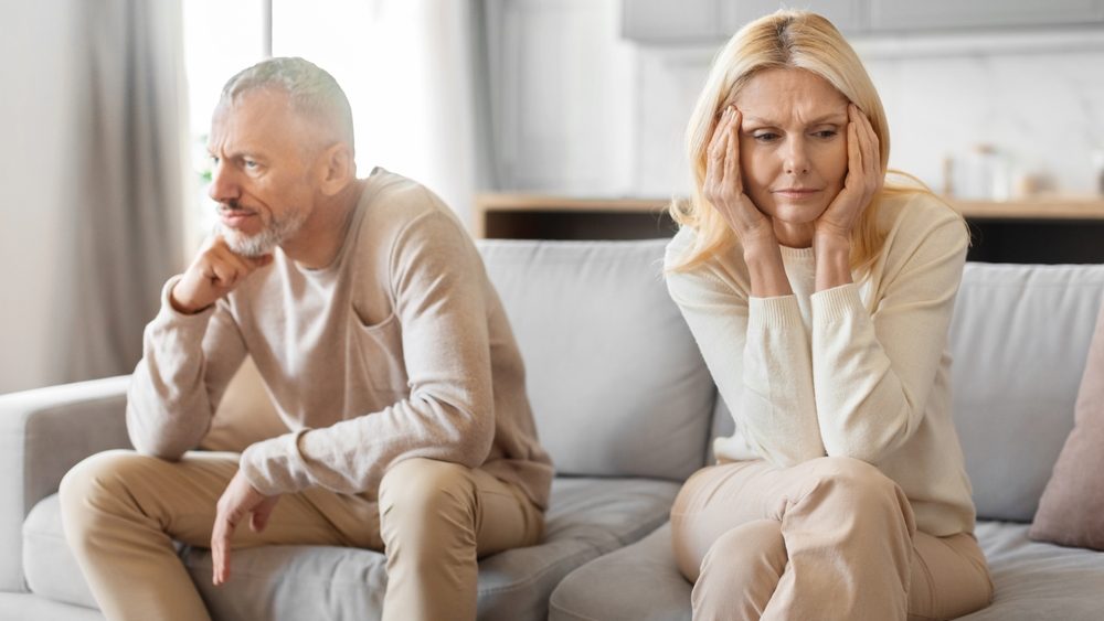A middle-aged man and woman sit apart on a couch, seemingly lost in their own deep thoughts