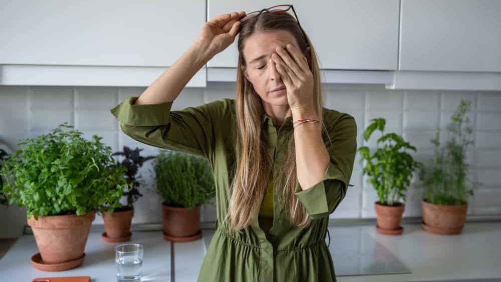 Sleepy woman suffering from chronic fatigue rubbing eyes on kitchen at home. Tired middle aged female feeling bad from lack of rest, headache, lack of energy after party, sleepless night. 