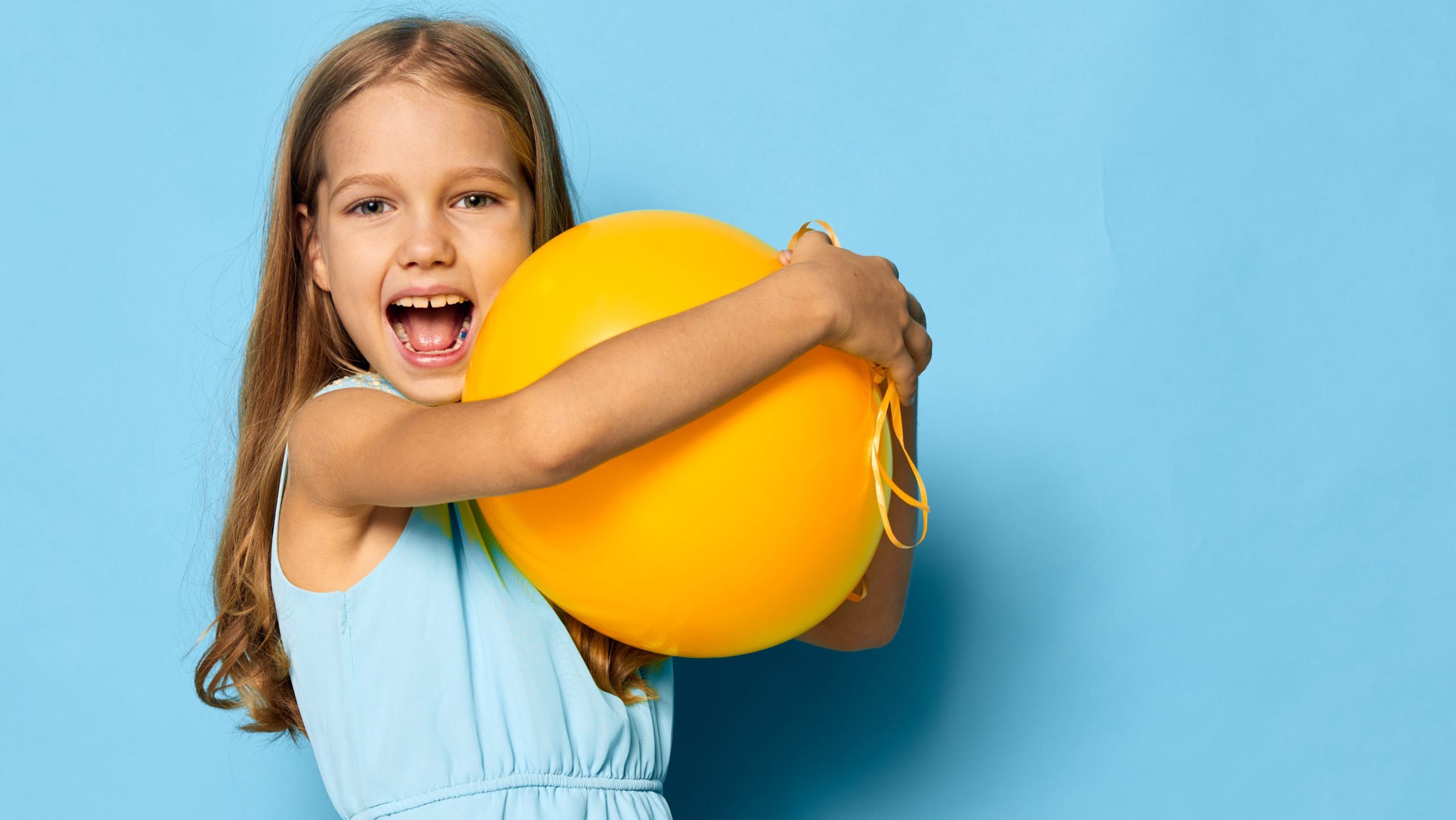 Joyful little girl in a blue dress holding a vibrant yellow balloon against a matching blue background