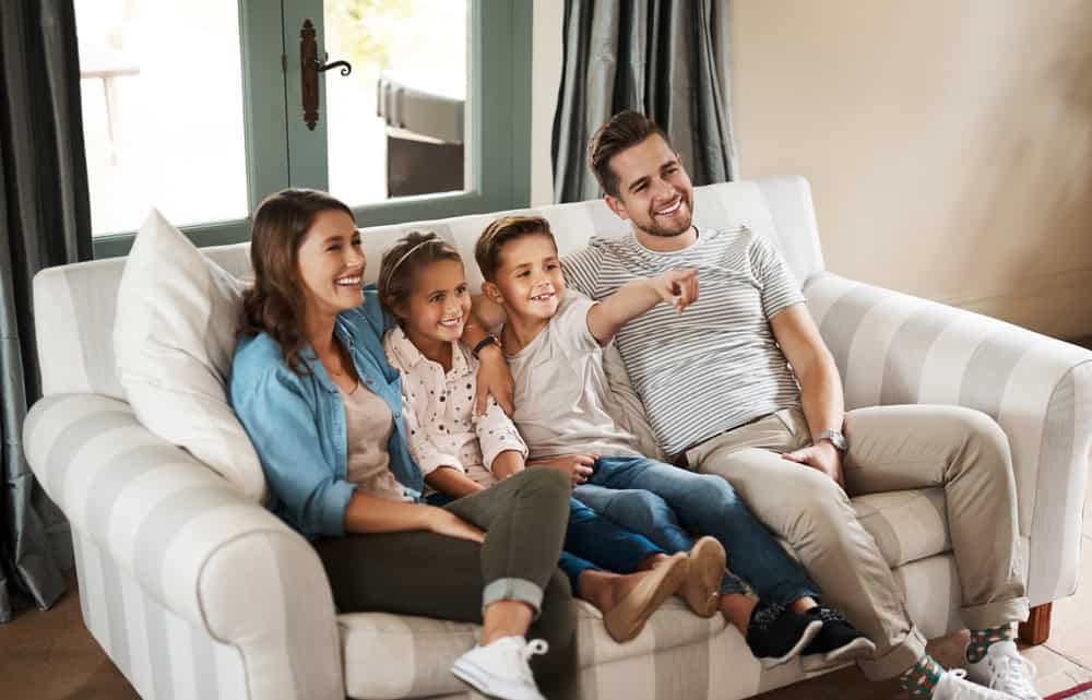 Parents, kids and watching tv on sofa with smile, point and relax together for bonding in family house. Father, mother and children with connection, happy and streaming with film, cartoon or movies
