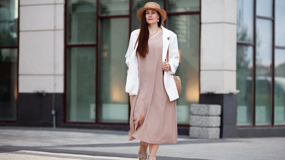 Stylish Plus size woman walks confidently outdoors in urban city, wearing beige dress, white blazer, wide brimmed hat, woven handbag. Model with long brown hair worn loose, calm, composed expression