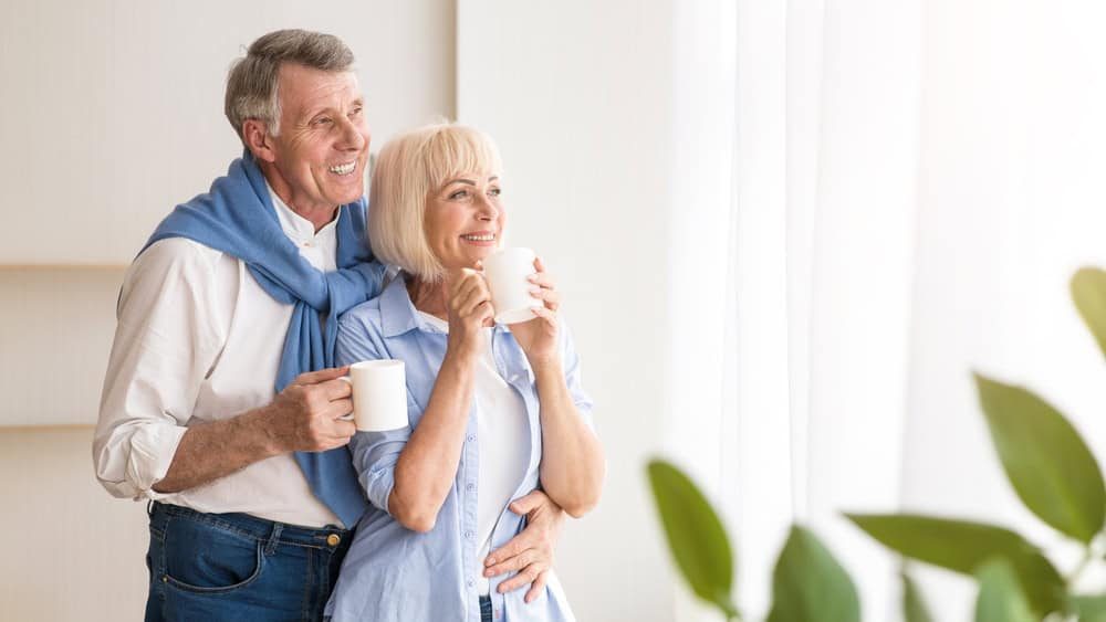 Happy senior couple drinking tea near window and embracing, free space