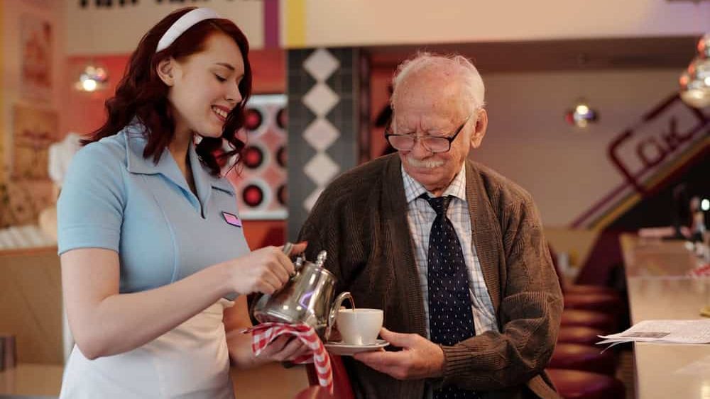 Smiling waitress serving coffee to elderly man seated at retro diner counter. Interaction between young waitress and elderly customer creating warm, nostalgic atmosphere
