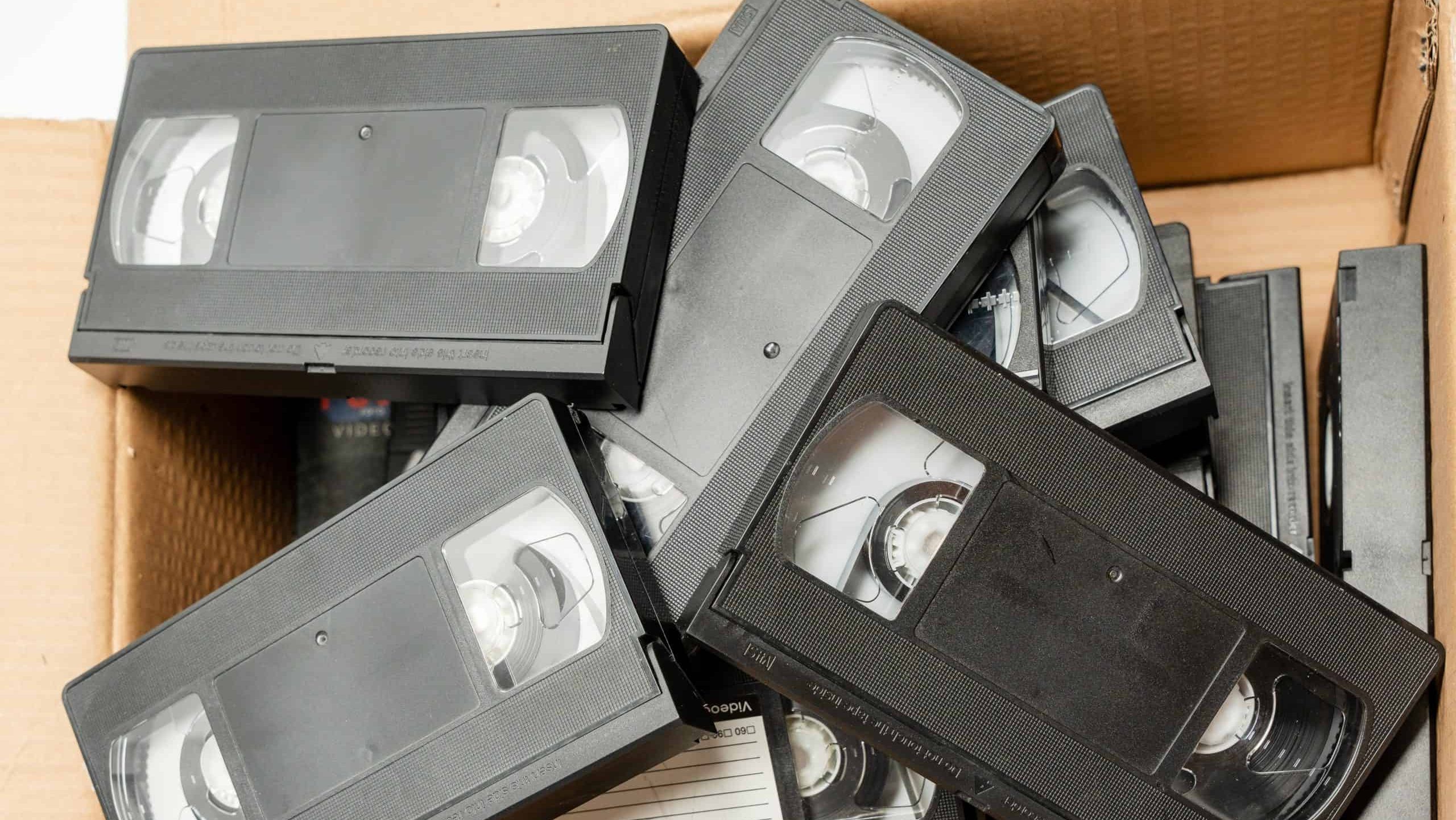A box filled with a large number of black VHS tapes. The tapes are old and worn, and they are stacked on top of each other in the box