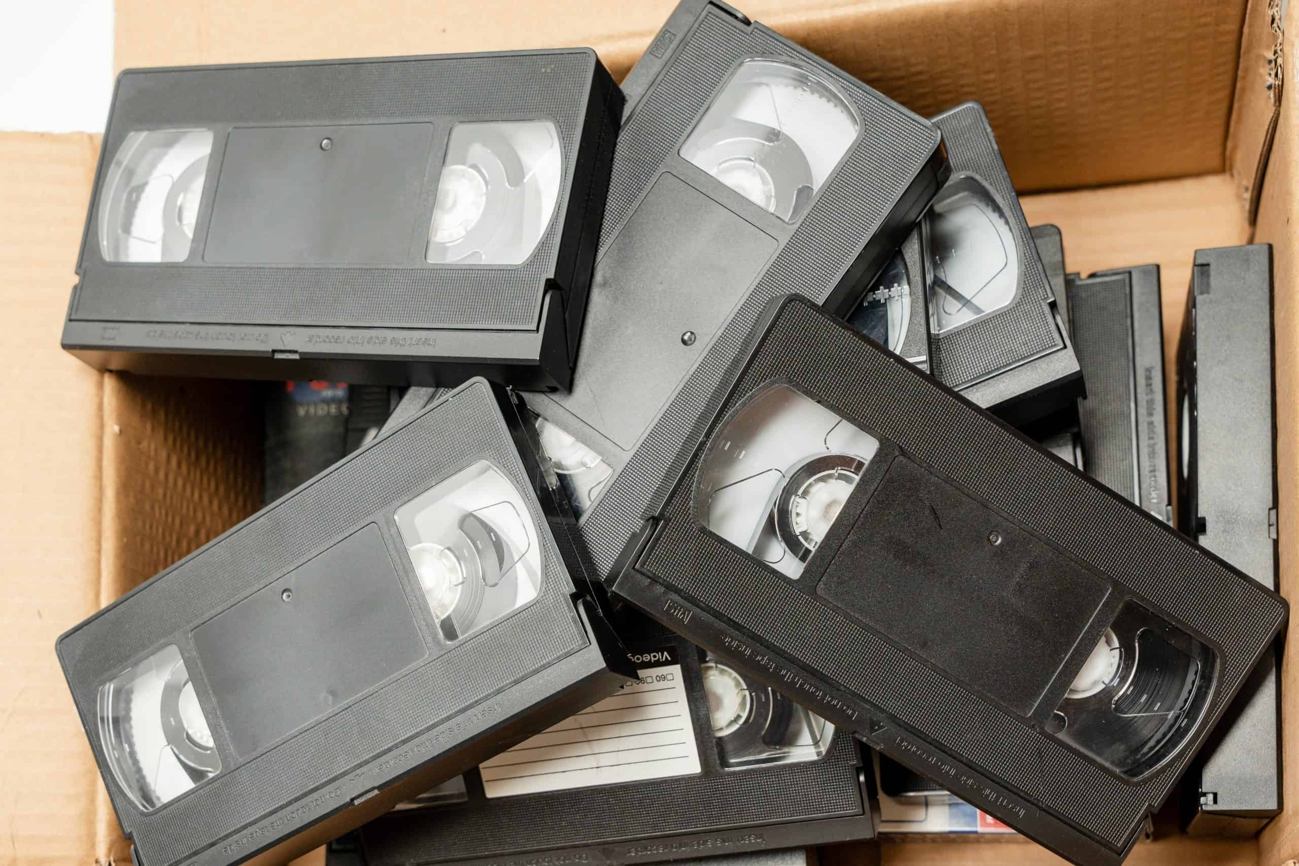 A box filled with a large number of black VHS tapes. The tapes are old and worn, and they are stacked on top of each other in the box