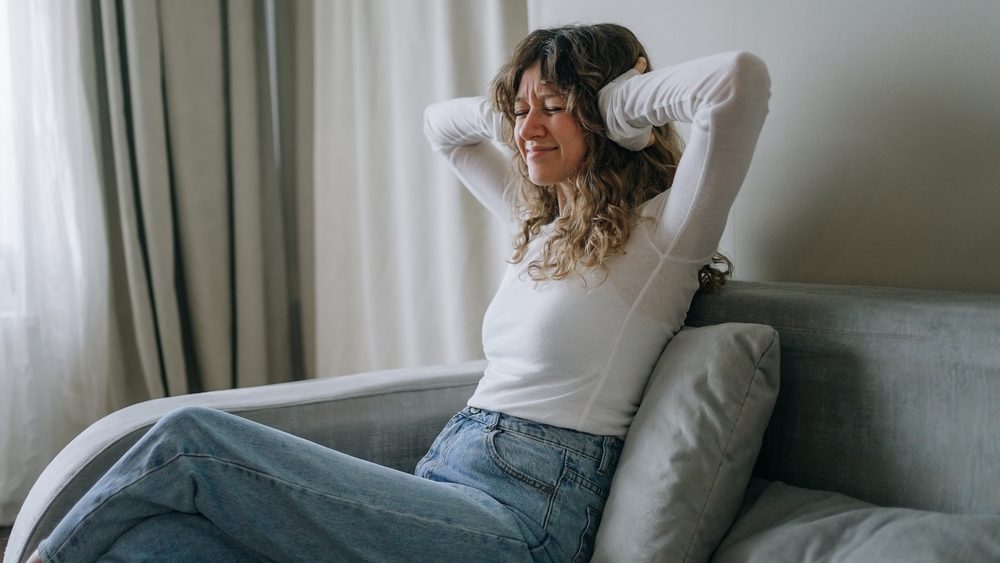 Stressed woman on sofa covering ears from loud noise, feeling annoyed and frustrated in living room