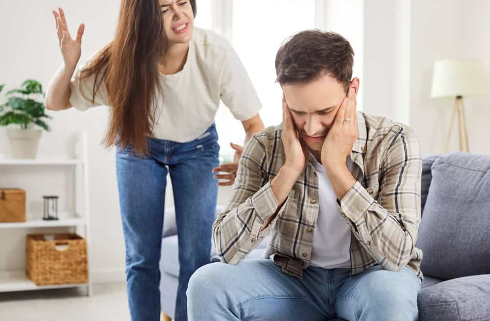 Young family couple having problems in relationships. Angry offended wife standing and shouting at her husband in quarrel, with frustrated stressed man sitting on sofa, closing his ears with his hands