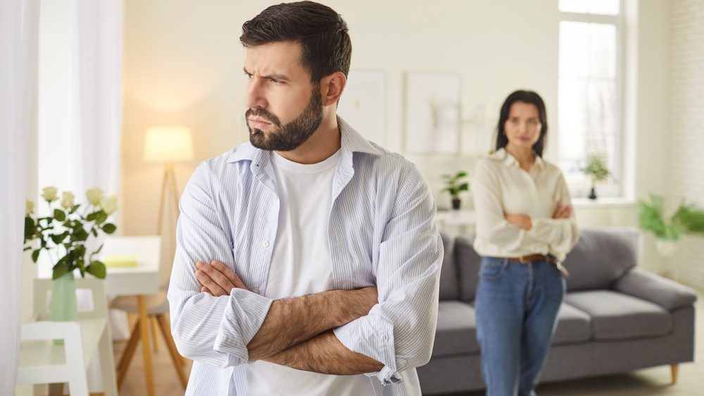 Portrait of young frustrated upset husband standing with his arms crossed and ignoring his wife. Upset young family couple having relationship problems, standing in silence in room after quarrel.
