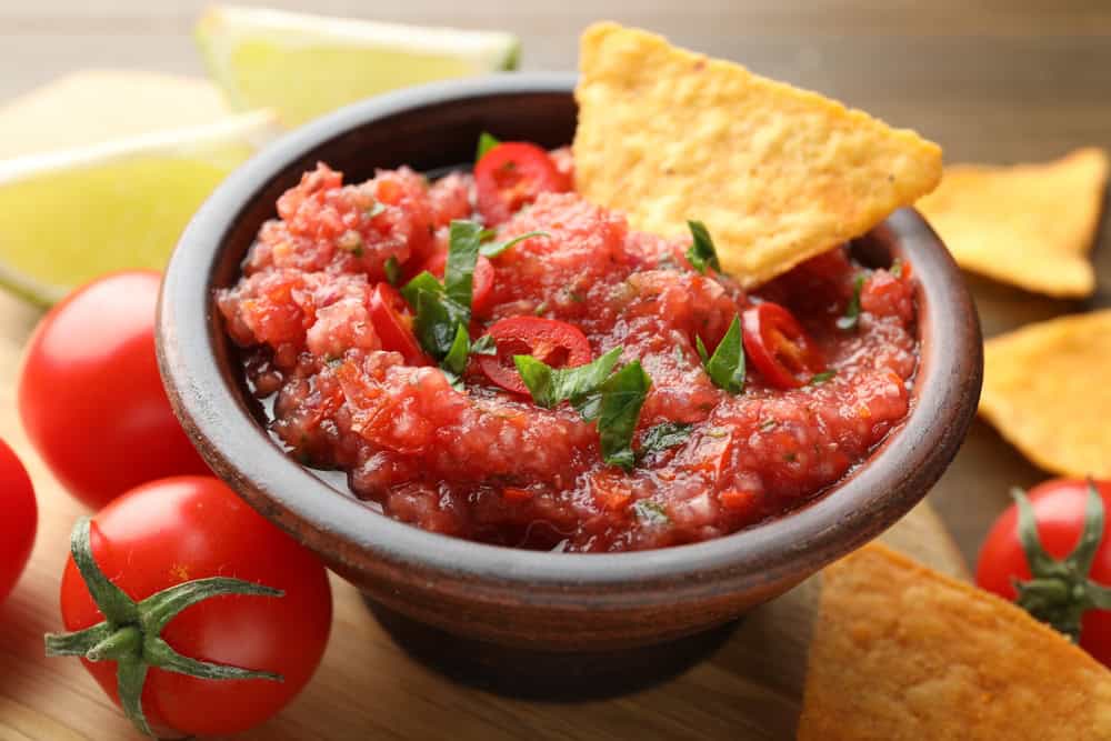 Spicy salsa sauce in bowl, nachos and tomatoes on table, closeup