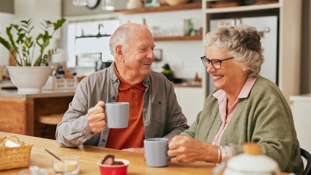 Smiling old couple sitting at table and sharing coffee in their cozy kitchen. Elder man and retired woman laughing together while enjoying a cup of coffee indoors. Relaxed aged couple drinking a tea.