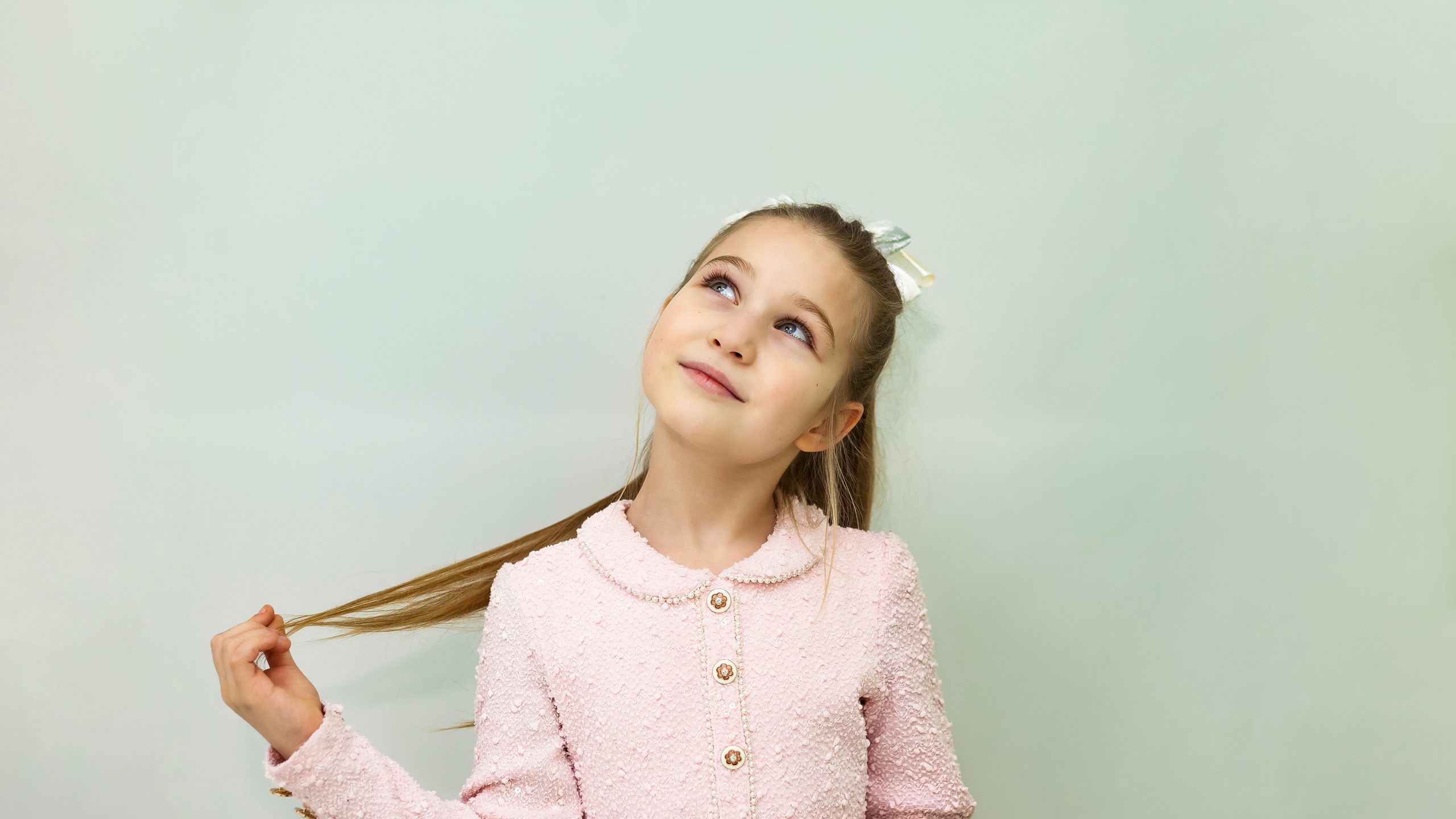 An eight-year-old girl model stands in a studio with a soft pastel backdrop, playfully twirling her hair. She wears a stylish light pink outfit, giving an impression of innocence and charm.