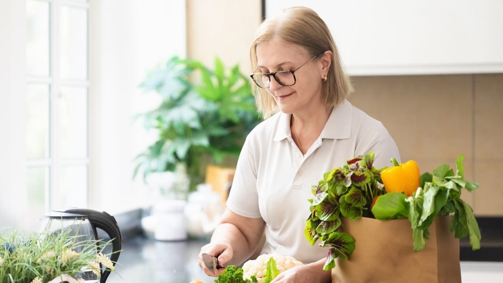 Senior woman holding a paper bag with vegetables. Healthy nutrition in retirement age. Zero waste home. Female in sunny kitchen making salad. Retired woman shopping. Fresh green lettuce, pepper bell.