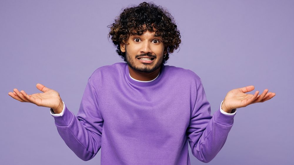 Young sad upset Indian man he wear violet sweatshirt casual clothes shrugging shoulders looking puzzled spread hands isolated on plain pastel light purple background studio portrait. Lifestyle concept