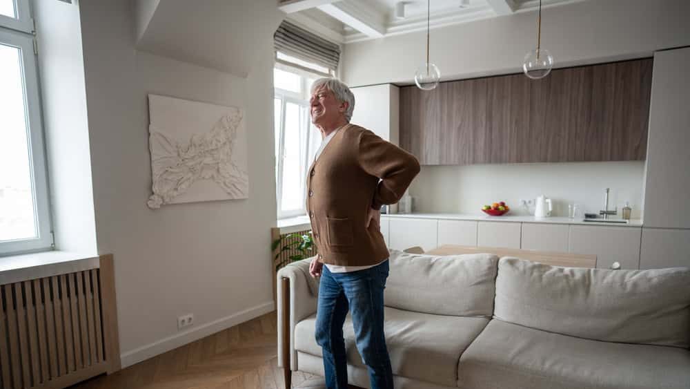 Grimacing senior man in pain winces while straightening hunched posture after standing up from couch, gripping aching back. Degenerative disease, nerve compression, kidney discomfort, limited mobility