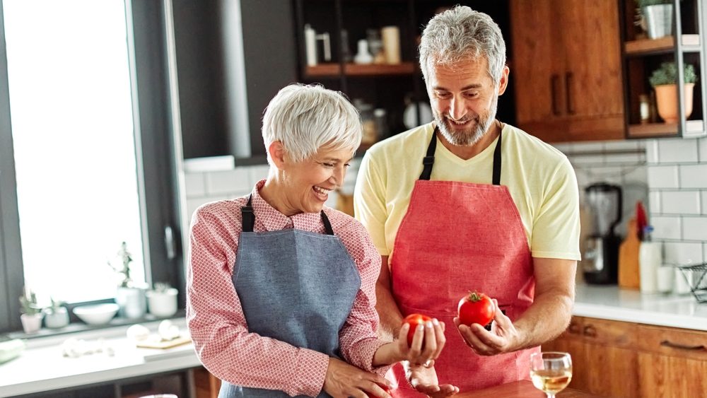 Portrait of happy senior mid aged mature couple prepering meal with fresh vegatebles in kitchen at hpme