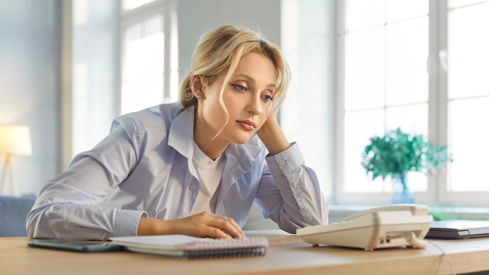 Tired young woman sitting at desk, leaning on hand and staring at landline phone. Exhausted anxious company employee waiting for important phone call, feeling stressed and tense while expecting news.