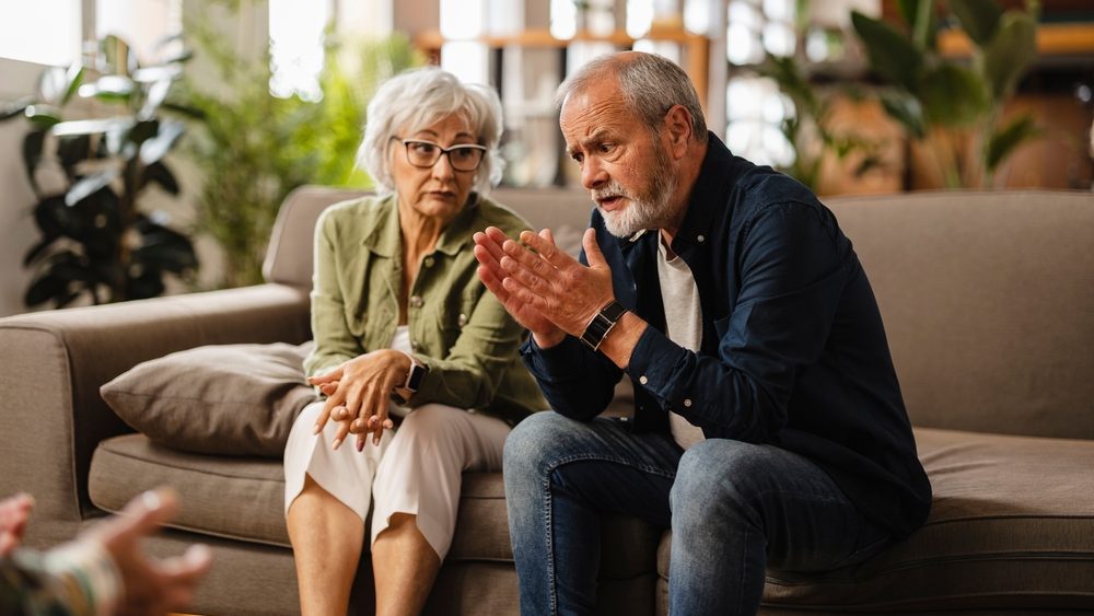 Senior couple sitting on a sofa, discussing their problems with a psychologist during a therapy session at home, sharing concerns and seeking guidance for their relationship issues