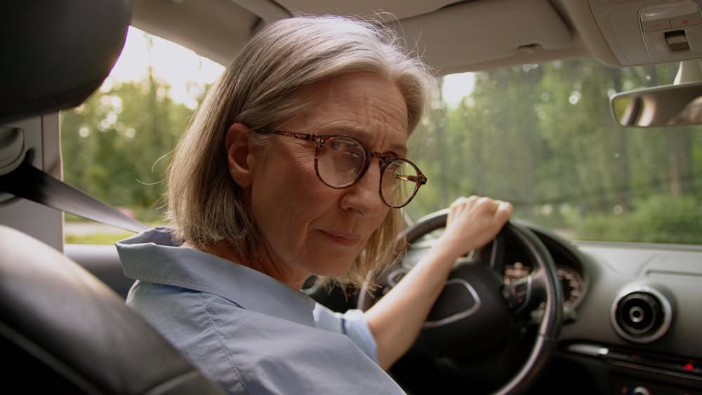 Displeased confused Caucasian aged woman senior female taxi driver mature car auto owner drive automobile steering wheel looking back at camera passenger seat raise eyebrow expression confusion indoor
