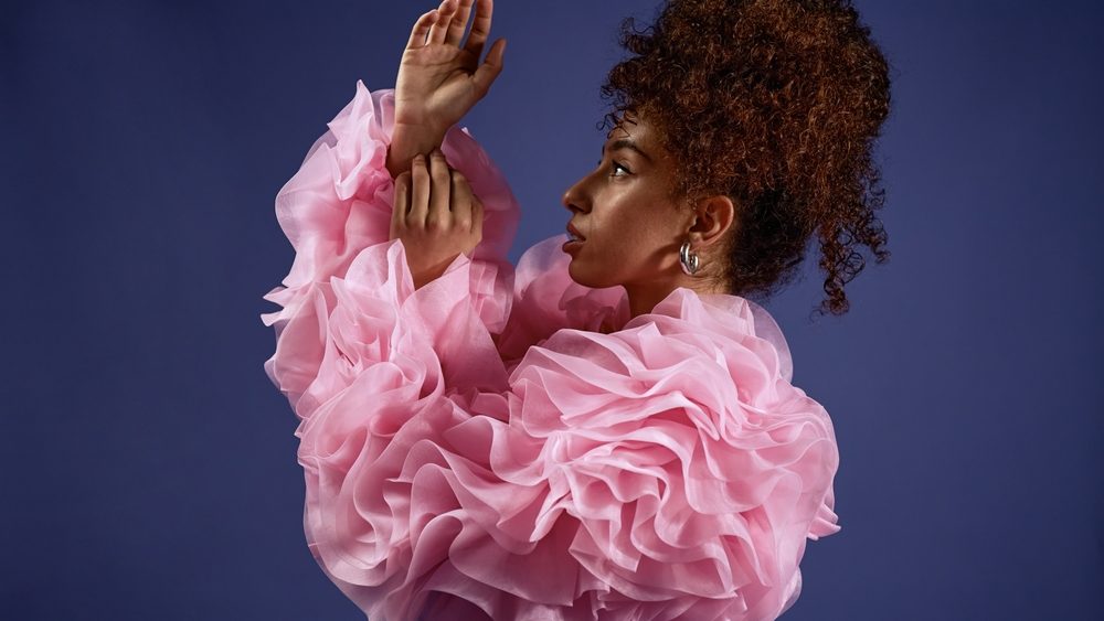 Vertical shot of young adult multiethnic woman posing in profile with arms raised, wearing voluminous ruffled top, curly hair styled up, participating in fashion photoshoot against plain background