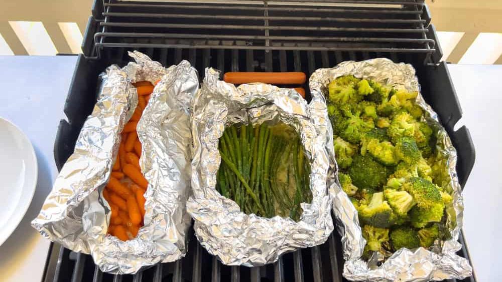 Top view of carrots, green beans, and broccoli in foil packets on a black barbecue grill. The food is arranged side by side and illuminated by natural sunlight.