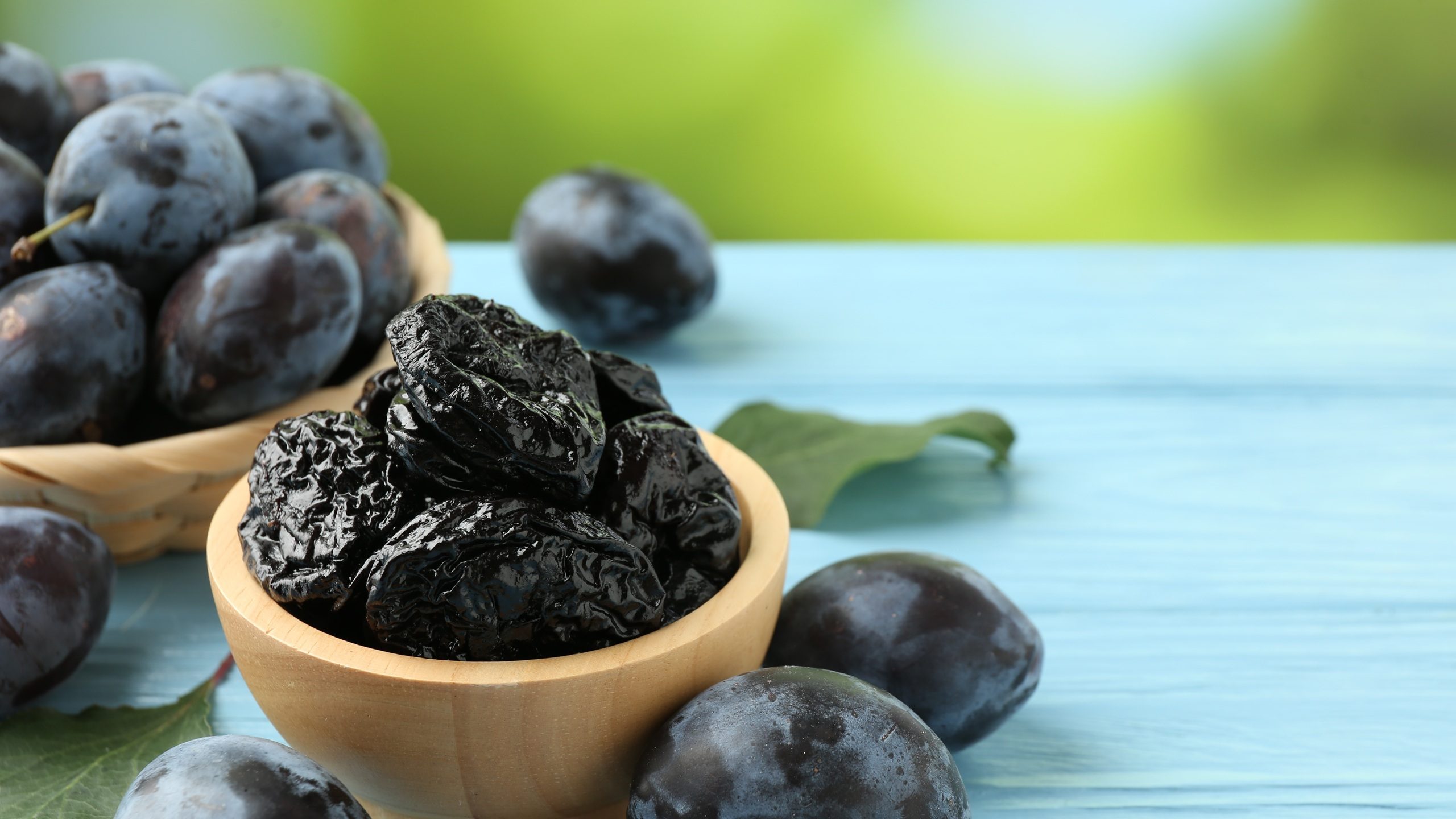 Dried prunes in bowl, leaves and fresh plums on light blue wooden table, closeup. Space for text