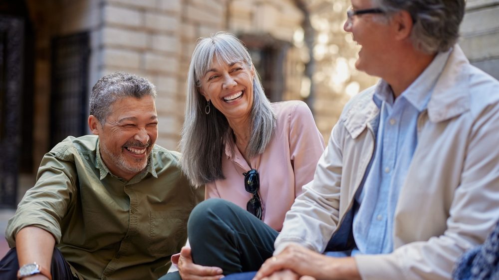 Older friends laugh together while sitting outside in the city. Happy seniors enjoy a funny moment during a walk. Mature women and old men smile and talk sitting on the steps of a square.