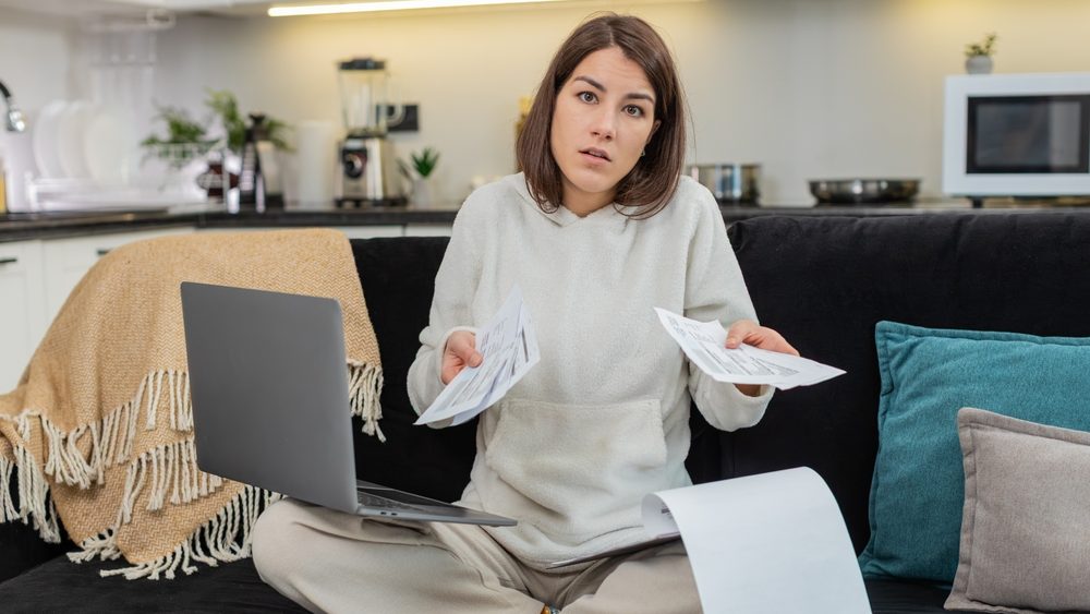 Young woman at home struggles to file finance papers on laptop sees bills feels confused and tense. Caucasian girl on sofa looks at camera asks about charges opens support chat and plans steps later