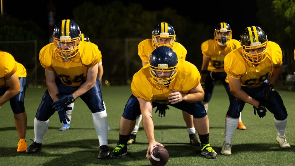 American football team on the field, players in offensive line positions, preparing to snap the ball during a nighttime game