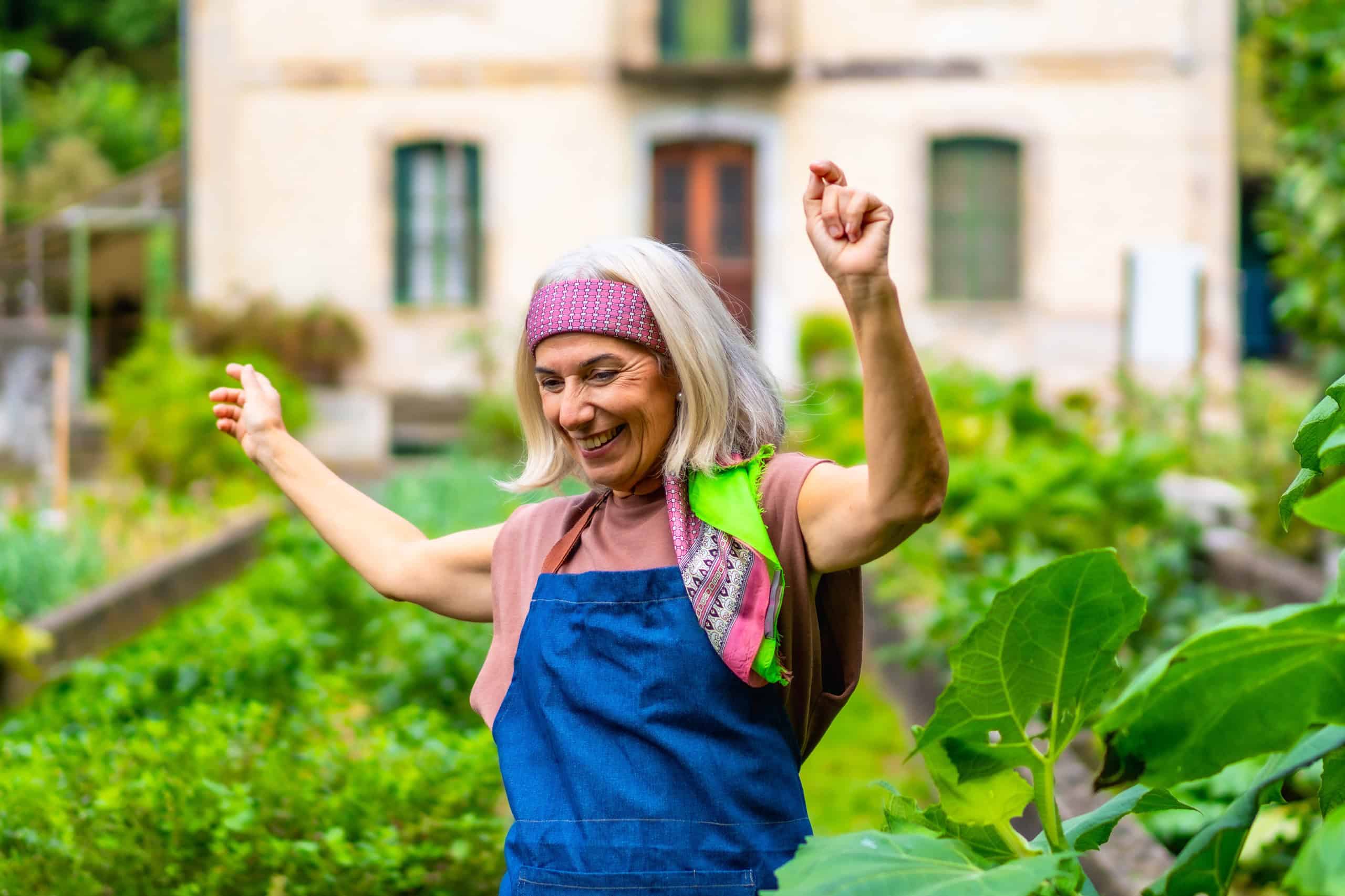 Happy senior woman dancing with arms raised in her home garden, celebrating an active and joyful retirement while cultivating fresh vegetables outdoors