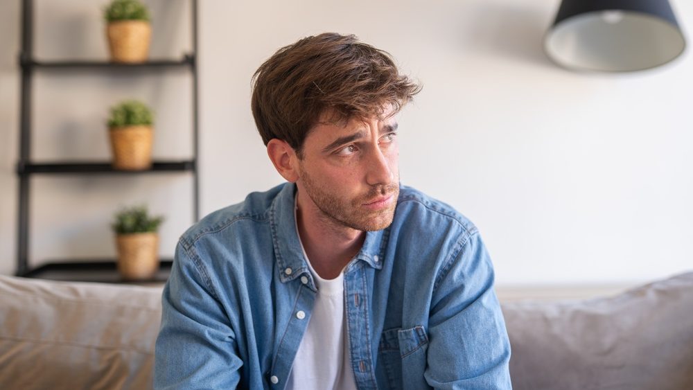 An upset millennial man sits alone on a sofa, thinking about personal problems and feeling anxious and disconnected in a silent home environment