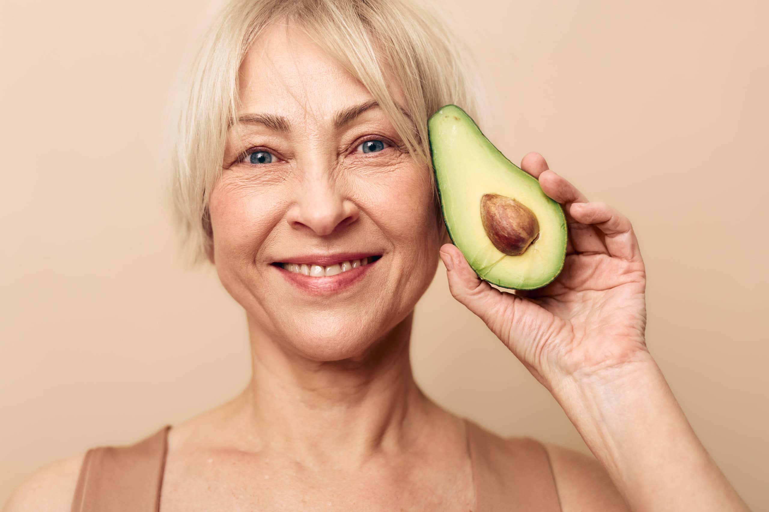 Smiling mature woman with short blonde hair holds fresh avocado half near face on beige background. Concept for healthy lifestyle, nutrition, natural beauty, and wellness at any age.