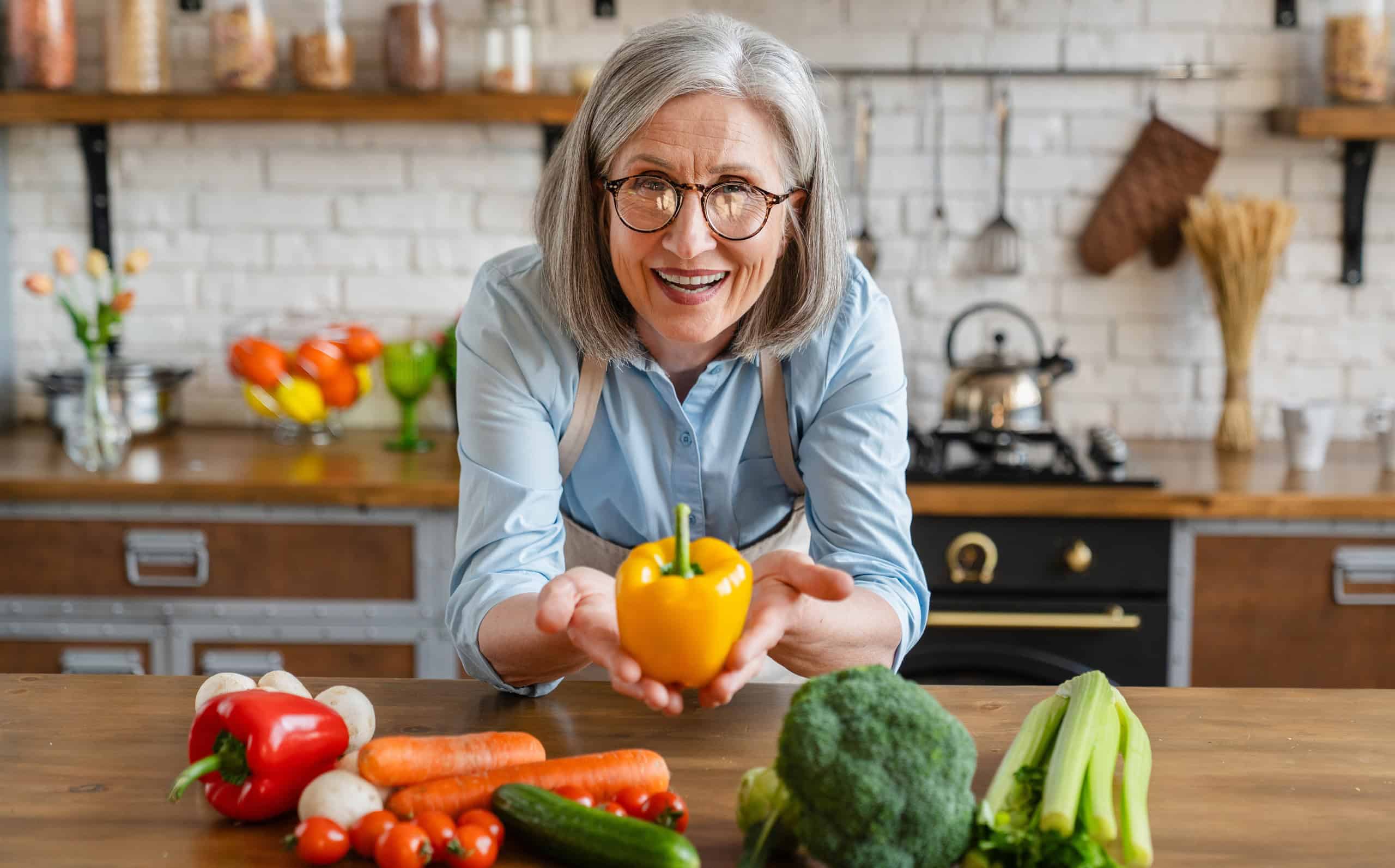 Woman holding fresh sweet yellow pepper above kitchen table with cutting board and healthy vegetables. Vitamin and fiber source. Dieting and healthy eating habits, slimming, nutrition concept