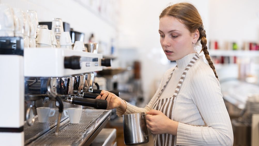 Teen girl coffee shop employee at work, selling coffee, cocoa and cocktails, uses coffee machine. Public catering place, location for coffee break.