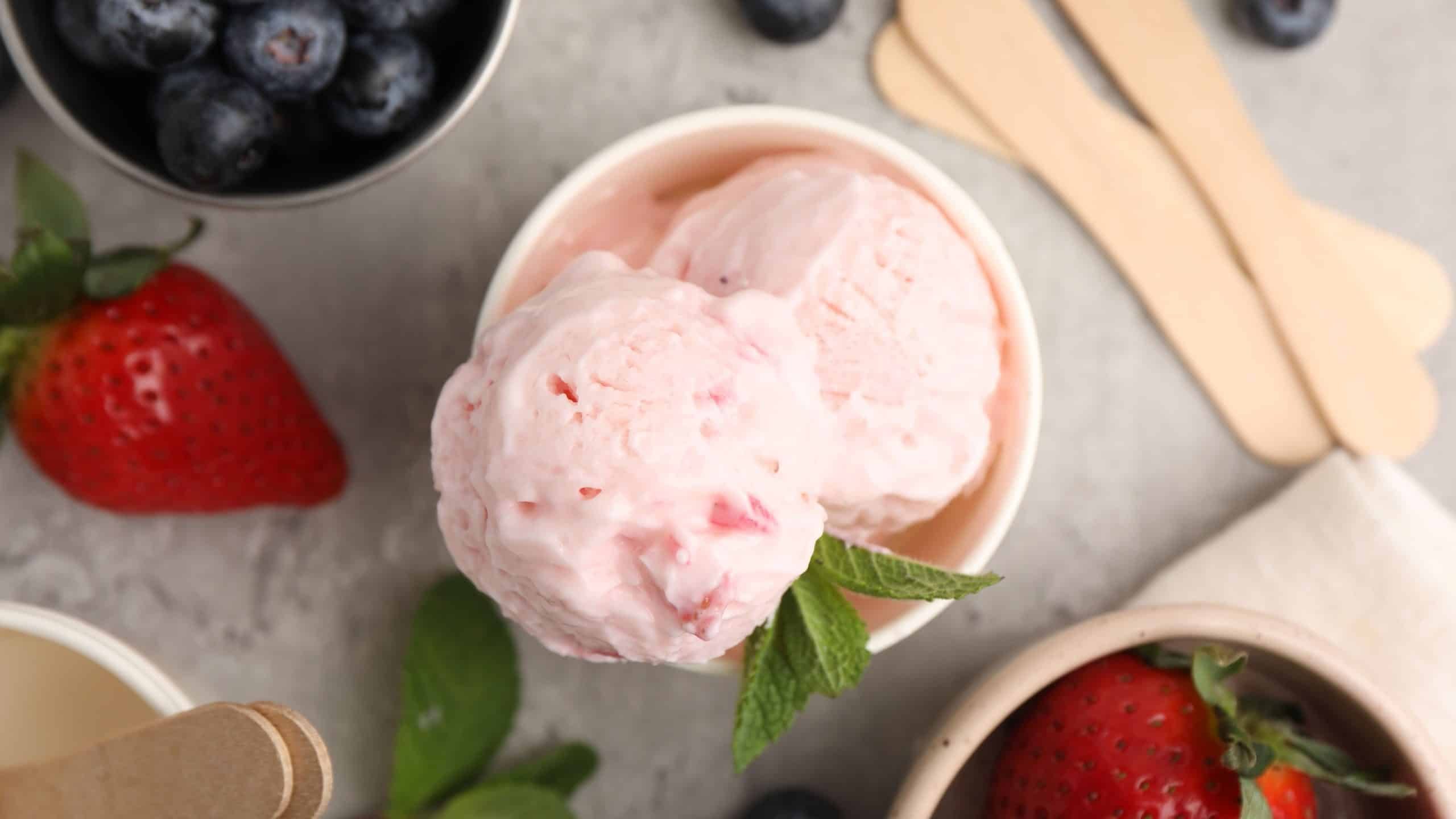 Tasty frozen yogurt in paper cup, berries and mint leaves on grey table, flat lay