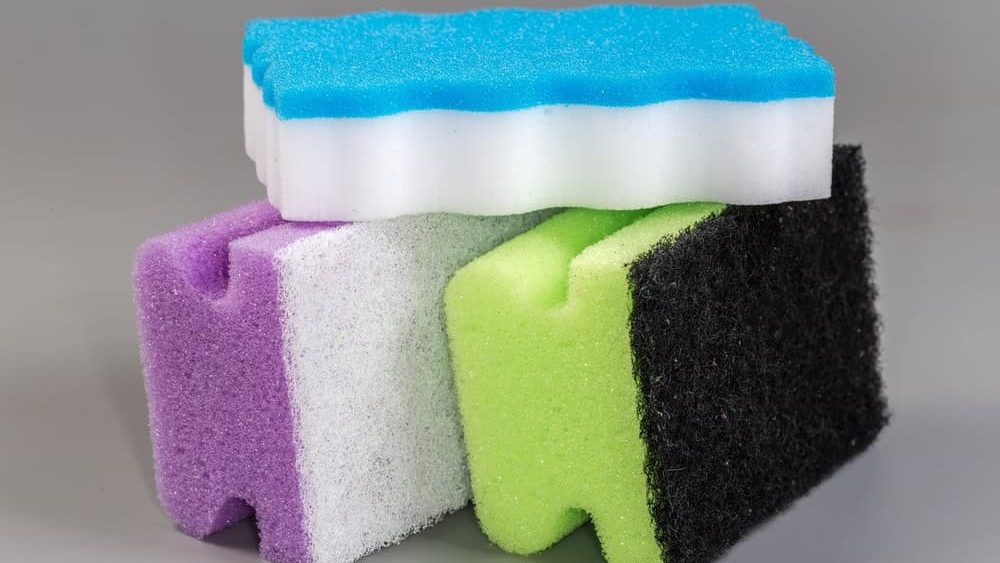 Synthetic multicolored polyurethane cleaning sponges, two with abrasive fiber layers located at bottom and one sponge with white melamine abrasive layer atop on a gray background