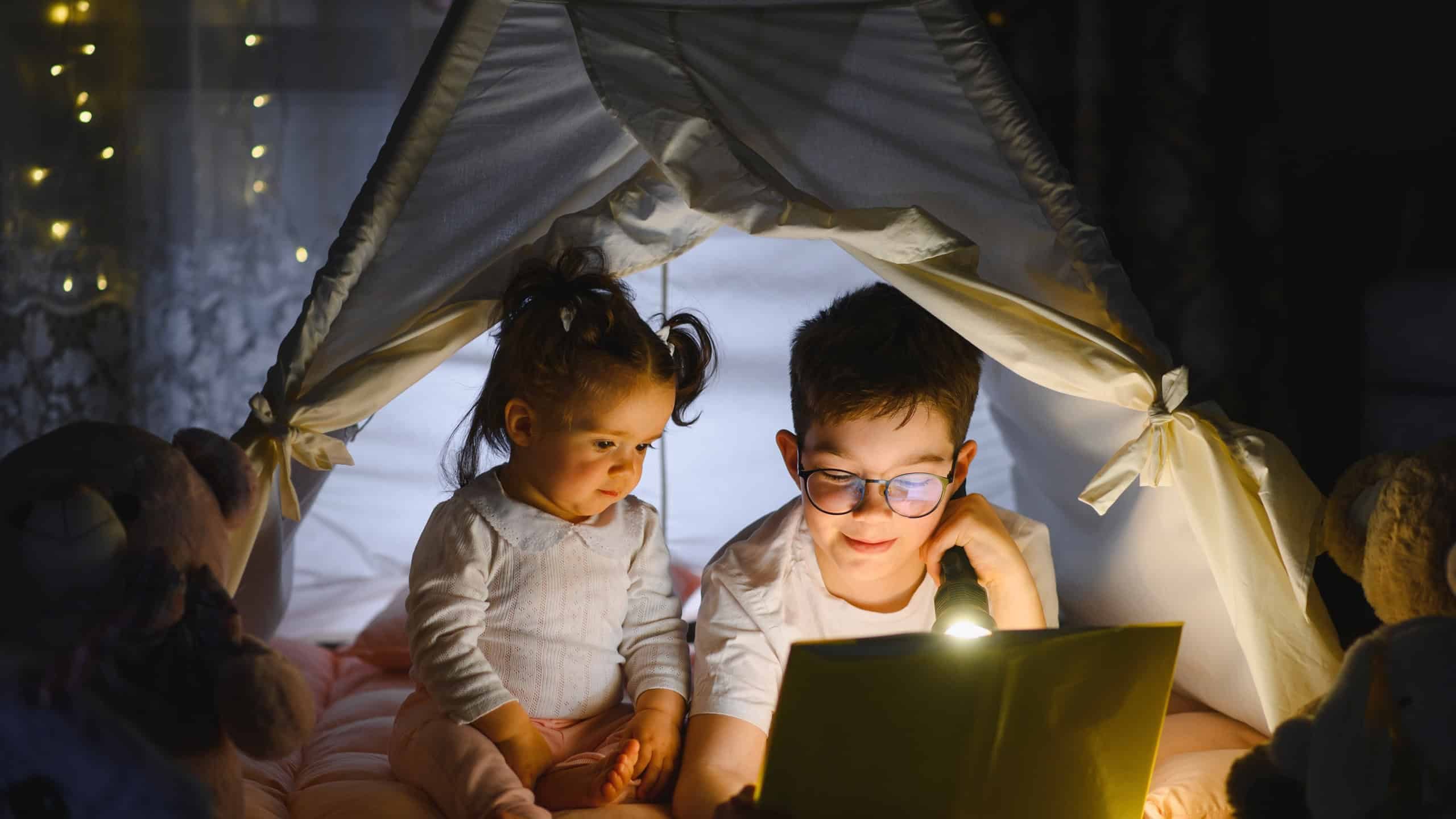 Young siblings enjoying storytelling in a dimly lit tent, illuminating a book with a flashlight at night