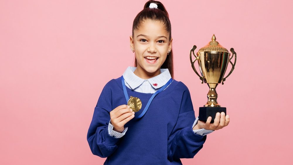 Little kid girl 8-9 years old she wears blue shirt golden medal hold in hand champion cup have fun isolated on pink color background child studio portrait. Mother's Day love family lifestyle concept