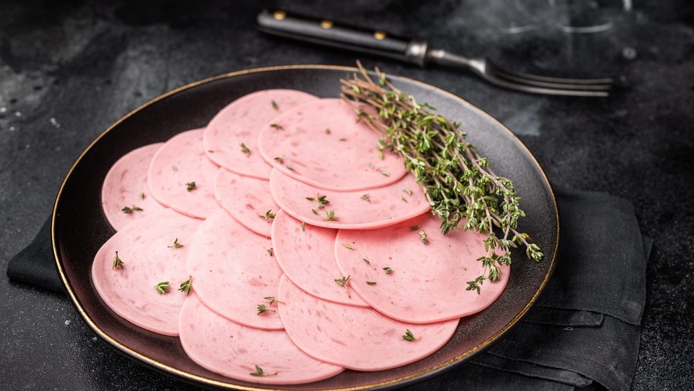 Sliced pink lunch meat (bologna mortadella) with fresh thyme on a black plate. Delicious cold cut and charcuterie concept on a dark background