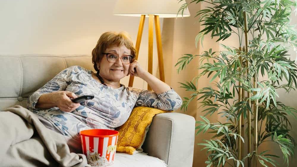 Smiling senior woman enjoying an evening at home, sitting on a sofa with popcorn and a tv remote