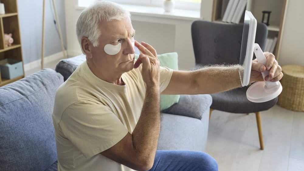 Senior man skincare with patches and mirror. He applies under eye pads at home, checking his face in a handheld mirror on the sofa to promote grooming beauty and anti aging treatment. Concept of male