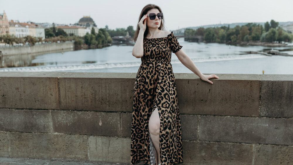 Portrait of one beautiful young Caucasian girl, brunette in sunglasses and in an animal print dress, standing on the Charles Bridge, looking to the side leaning her hand on the stone railing on a summer day in Prague, side view, general plan with copy space on the left. City walk concept.
