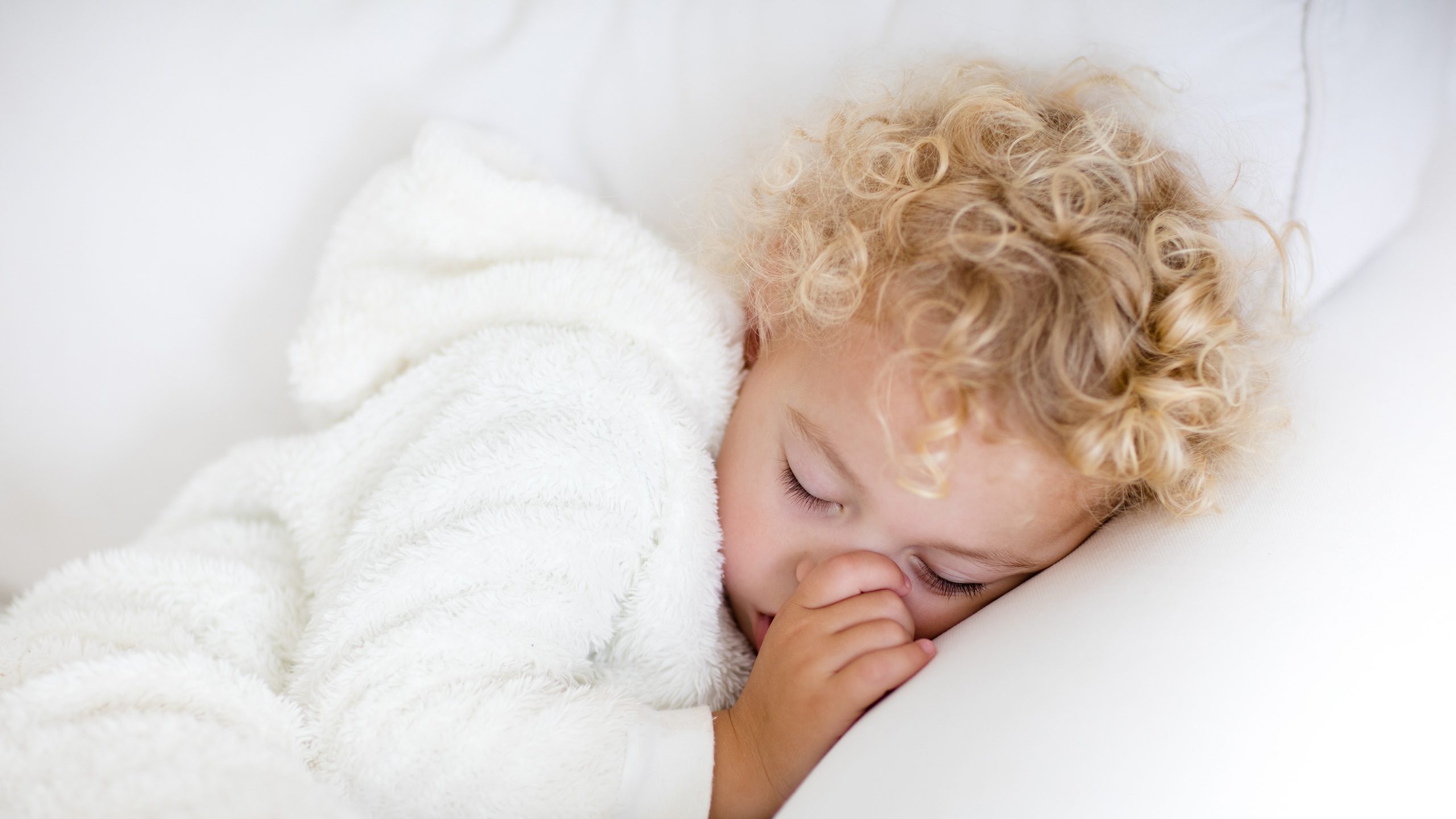 Cute blond curly little boy sleeping on white couch. Tired toddler taking a nap wearing warm white pajamas sucking his thumb.