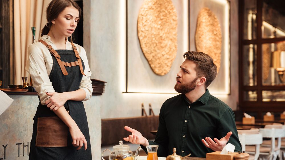 Image of angry bearded young man sitting in cafe. Looking at waiter.